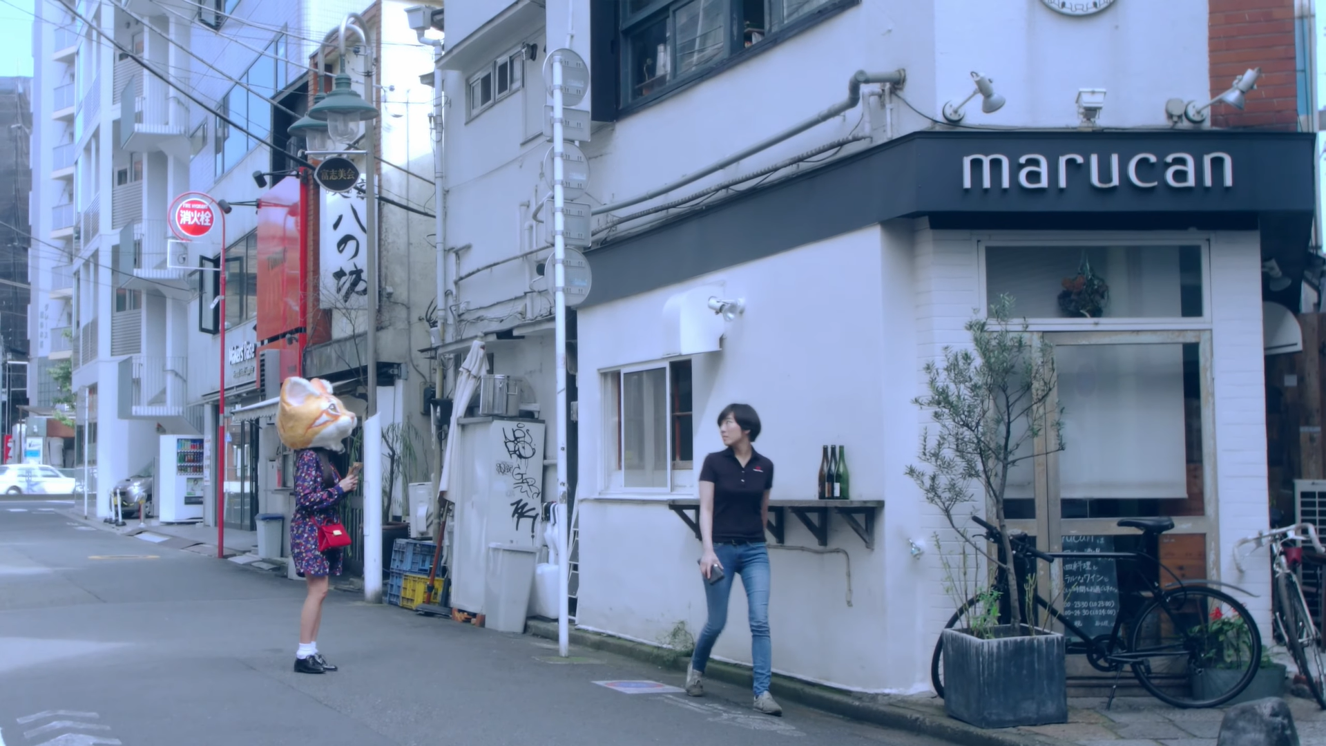 Hyunjin standing on the street, looking into the window of the building in front of her. She is wearing a large orange cat head.